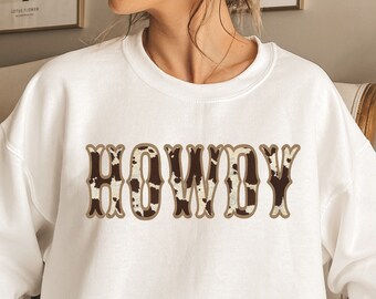 Op de afbeelding: Wit sweatshirt met een bruin en wit koeienprintontwerp met de tekst "HOWDY" in een vintage lettertype.