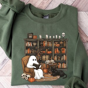 May include: A green sweatshirt with a graphic of a ghost reading a book in a chair next to a bookshelf filled with books. There are pumpkins, skulls, and candles on the shelf.