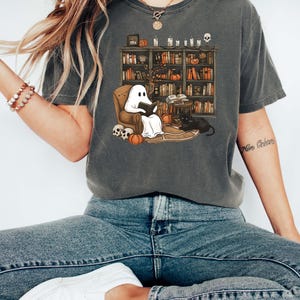May include: A gray t-shirt with a graphic of a ghost reading a book in a chair in front of a bookshelf. The bookshelf is filled with books, candles, and other Halloween decorations. The text "More Brains" is visible on the right side of the shirt.