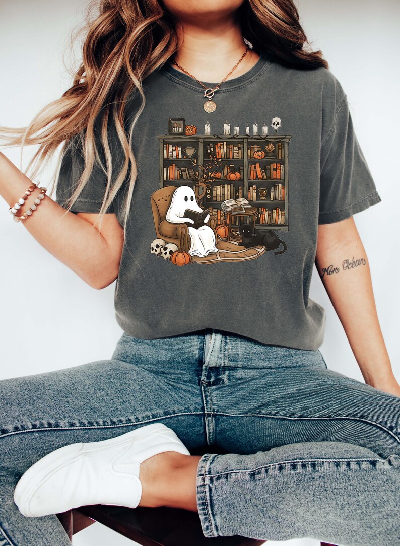 May include: A gray t-shirt with a graphic of a ghost reading a book in a chair in front of a bookshelf. The bookshelf is filled with books, candles, and other Halloween decorations. The text "More Brains" is visible on the right side of the shirt.