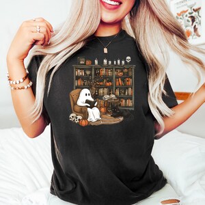 May include: Black t-shirt with a graphic of a ghost reading a book in a chair next to a bookshelf and a black cat. The bookshelf is filled with books and has a skull on top.