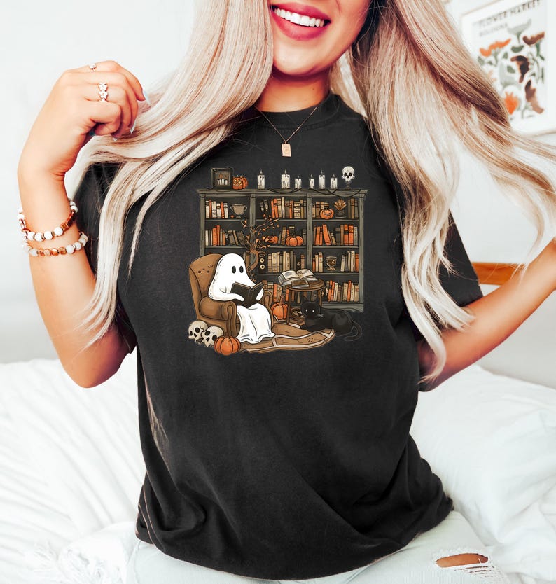 May include: Black t-shirt with a graphic of a ghost reading a book in a chair next to a bookshelf and a black cat. The bookshelf is filled with books and has a skull on top.