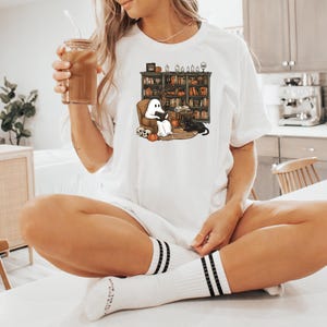 May include: A white t-shirt with a graphic of a ghost and a black cat reading in a library. The ghost is wearing a white sheet and has a book in its hands. The cat is sitting on a book and has a book in its paws. The library is filled with bookshelves and has a fireplace in the background.