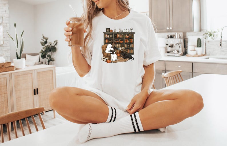 May include: A white t-shirt with a graphic of a ghost and a black cat reading in a library. The ghost is wearing a white sheet and has a book in its hands. The cat is sitting on a book and has a book in its paws. The library is filled with bookshelves and has a fireplace in the background.