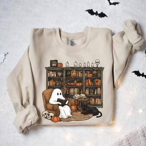 May include: A beige sweatshirt with a graphic print of a ghost reading a book in a chair next to a bookshelf filled with books and candles. The ghost is wearing a white sheet and has a black book in its hands. There is a black cat sitting on the floor next to the chair.