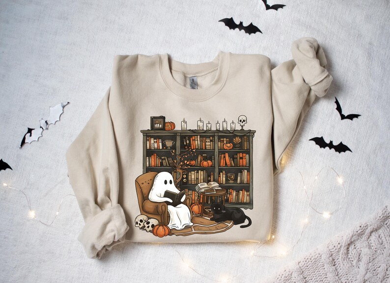 May include: A beige sweatshirt with a graphic print of a ghost reading a book in a chair next to a bookshelf filled with books and candles. The ghost is wearing a white sheet and has a black book in its hands. There is a black cat sitting on the floor next to the chair.