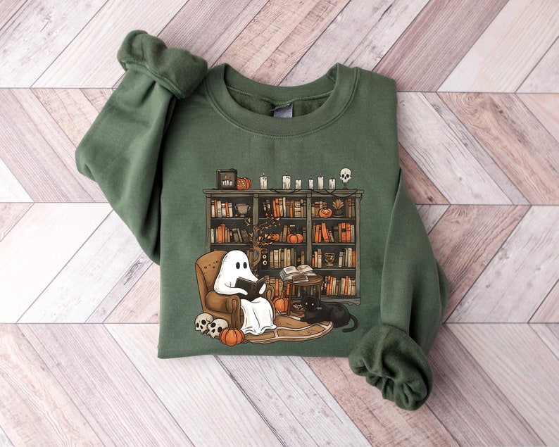 May include: A green sweatshirt with a graphic of a ghost reading a book in a chair next to a bookshelf filled with books. There are pumpkins, skulls, and candles on the shelf.