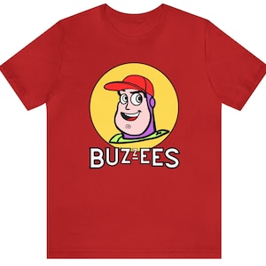 May include: Red t-shirt with a cartoon image of Buzz Lightyear from Toy Story wearing a red hat and a yellow circle around his head. The text "BUZZ-EES" is printed below the image.