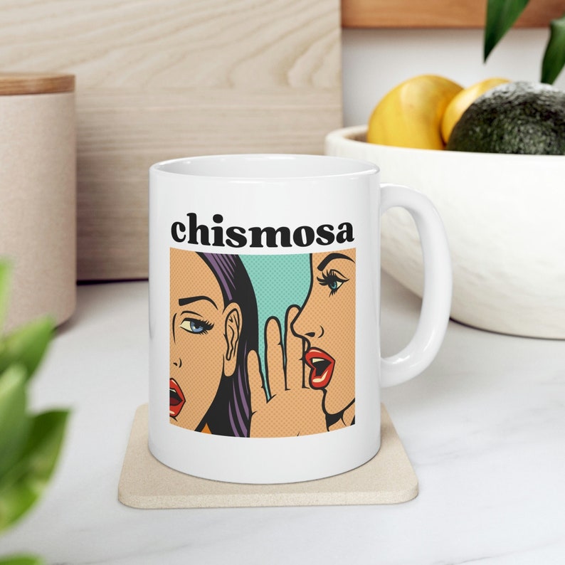 May include: White ceramic mug featuring a black and white illustration of two women whispering. The text "chismosa" is printed in black above the illustration.