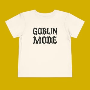 Puede incluir: Camiseta de color crema con las palabras "GOBLIN MODE" impresas en una fuente gótica. El texto está centrado en la parte delantera de la camiseta. La camiseta tiene mangas cortas y cuello redondo.
