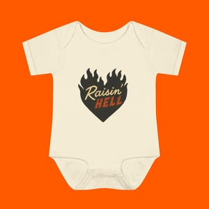 Puede incluir: Un body blanco para bebé con un diseño de llama en forma de corazón negro y el texto "Raisin' Hell" en naranja y amarillo.