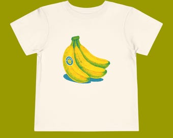 Op de afbeelding: Off-white peuter t-shirt met een afbeelding van drie gele bananen met groene stelen. Eén banaan heeft een blauwe sticker met een smiley en het woord "BANANA". De bananen werpen een blauwe schaduw.