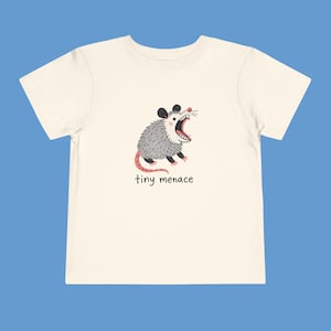 Op de afbeelding: Crèmekleurig peuter-T-shirt met een cartoonopossum. De opossum heeft grijze vacht, een roze staart en een open mond. De tekst "tiny menace" staat eronder. De achtergrond is blauw.
