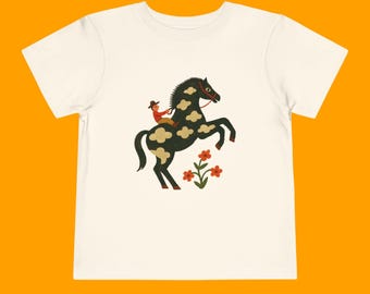 May include: White t-shirt with a black and white illustration of a person riding a horse with clouds on its body. The horse is rearing up and there are red flowers in the background.