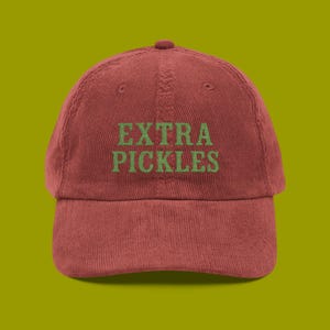 Op de afbeelding: Rode corduroy baseballpet met de woorden "EXTRA PICKLES" in groen geborduurd op de voorkant. De pet heeft een gebogen klep en een afgeronde bovenkant.