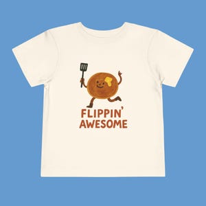 Può includere: T-shirt color crema per bambini piccoli con un personaggio di pancake dei cartoni animati con una spatola, che corre e sorride. Il testo "FLIPPIN' AWESOME" è stampato in marrone sotto il pancake. La maglietta ha maniche corte e girocollo.