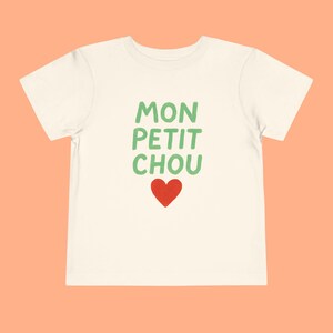May include: Off-white toddler t-shirt featuring the French phrase "MON PETIT CHOU" in green, with a red heart beneath. The shirt is made of a soft fabric, suitable for children.