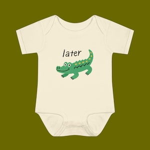 May include: Cream-colored baby onesie with a green alligator illustration and the word "later" above it. The alligator has yellow spots and a curved tail. The onesie has short sleeves and a simple design.