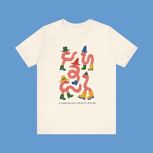 Könnte beinhalten: Cremefarbenes T-Shirt mit einer skurrilen Illustration von rosa Würmern, die Hüte und Stiefel in verschiedenen Farben tragen. Der Text unten lautet: "IF A WORM WORE BOOTS, HOW WOULD IT WEAR THEM?"