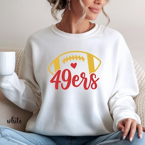 May include: White sweatshirt featuring a yellow and white football graphic with a red heart and the text "49ers" in red. The word "white" is printed in black at the bottom left. The sweatshirt is a casual style.