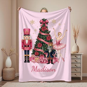 May include: A pink blanket featuring a Christmas tree, a nutcracker, a ballerina, and a mouse king. The tree is decorated with red poinsettias and a large bow. The blanket has the name "Madison" printed on it and gold snowflakes.