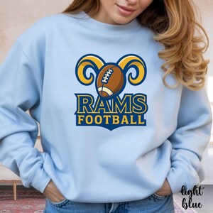 May include: Light blue sweatshirt featuring a football and ram horn graphic. The design includes a brown football, yellow ram horns, and the words "RAMS FOOTBALL" in blue and yellow. The sweatshirt is a light blue color.