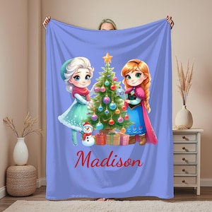 Puede incluir: Una manta lila con un &aacute;rbol de Navidad y dos personajes de dibujos animados. La manta tiene el nombre "Madison" en escritura roja. Los personajes visten ropa de invierno, y tambi&eacute;n se ven un mu&ntilde;eco de nieve y regalos envueltos.
