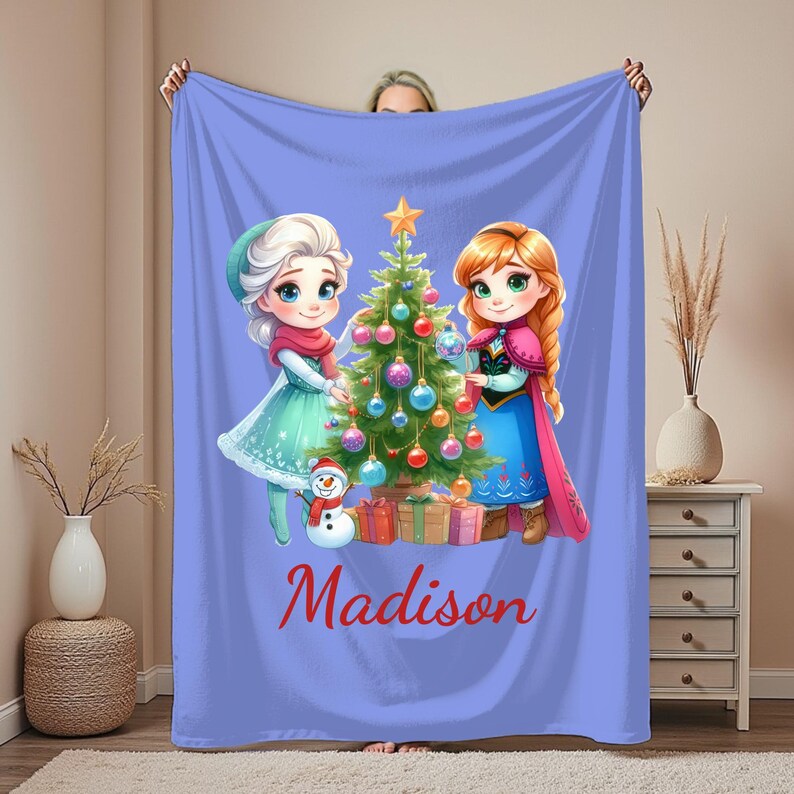 Puede incluir: Una manta lila con un &aacute;rbol de Navidad y dos personajes de dibujos animados. La manta tiene el nombre "Madison" en escritura roja. Los personajes visten ropa de invierno, y tambi&eacute;n se ven un mu&ntilde;eco de nieve y regalos envueltos.