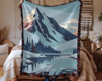 May include: A woven throw blanket featuring a mountain landscape design. The blanket displays a mountain range, a lake, and trees in shades of blue, gray, and brown. The edges are finished with a fringe in black, red, and brown.