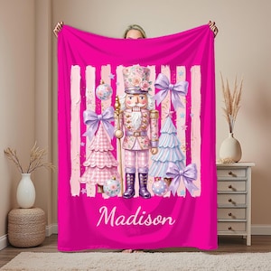 May include: A bright pink fleece blanket featuring a central nutcracker illustration surrounded by pink and white Christmas trees, bows, and ornaments. The design includes vertical brushstroke accents and the name Madison in white script lettering.