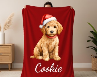 May include: A red blanket featuring a golden doodle puppy wearing a Santa hat and red collar. The puppy is sitting and the word "Cookie" is written in white script at the bottom of the blanket. The blanket is held up by a person.