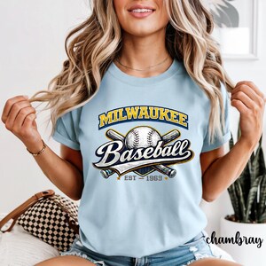 Puede incluir: Camiseta azul claro con el logo de Milwaukee Baseball. El diseño presenta una pelota de béisbol, bates cruzados y el texto "Milwaukee Baseball" y "EST - 1969". La camiseta tiene cuello redondo y mangas cortas, en un estilo vintage.