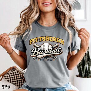 Puede incluir: Camiseta gris con un diseño de béisbol. El diseño incluye el texto "Pittsburgh Baseball" en amarillo y blanco, una pelota de béisbol y bates cruzados. También se incluye el texto "EST 1882".