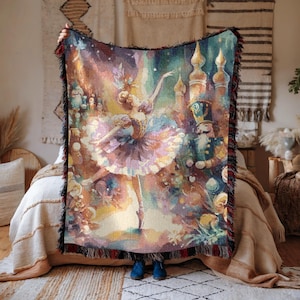 May include: A woven throw blanket featuring a ballet scene with a ballerina, a nutcracker, and castle turrets. The blanket has a colorful, painterly design with a fringed edge. The color palette includes pinks, blues, and golds.
