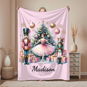 May include: A pink blanket featuring a ballerina, Christmas tree, nutcracker, and mouse king illustration. The blanket has a personalized name at the bottom. The design includes gold ornaments, snowflakes, and wrapped gifts.