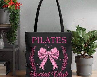 May include: Black tote bag with the words "PILATES Social Club" in pink script. A pink bow graphic is centered below the word "PILATES". The bag has black handles and is sitting on a wooden surface.