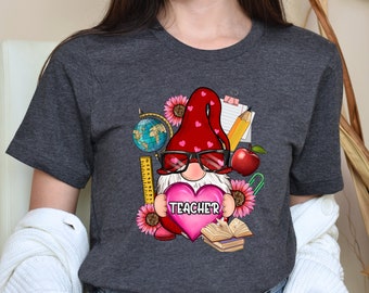 May include: Dark grey heather t-shirt featuring a graphic of a gnome wearing sunglasses and holding a heart that says "Teacher". The gnome is surrounded by school supplies such as a globe, ruler, pencil, apple, and books.