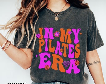 May include: A dark gray t-shirt with a retro-style graphic that says "In My Pilates Era" in pink and orange letters with stars and flowers.
