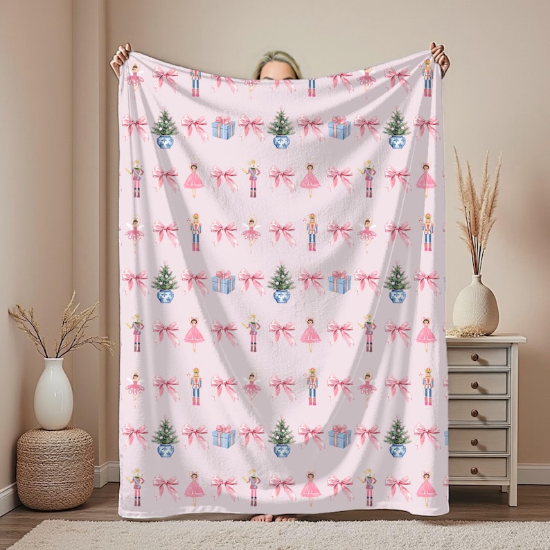Puede incluir: Una manta rosa con un patr&oacute;n repetido de &aacute;rboles de Navidad, cajas de regalo, lazos, bailarinas y cascanueces. La manta est&aacute; sujeta, mostrando el dise&ntilde;o. La manta est&aacute; hecha de un material suave.