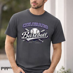 Puede incluir: Camiseta gris oscuro con un diseño de béisbol. El diseño incluye las palabras "Colorado Baseball" en blanco y morado, una pelota de béisbol y bates cruzados. También se incluye el texto "EST 1993".