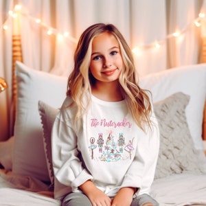 Puede incluir: Una sudadera blanca de manga larga con una ilustración rosa y azul de un árbol de Navidad con temática de Cascanueces. El texto "The Nutcracker" está impreso en rosa en la parte delantera de la sudadera.