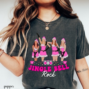 May include: A dark gray t-shirt with a pink graphic of four women wearing Santa hats and dresses. The graphic includes a boombox and the text "Jingle Bell Rock".