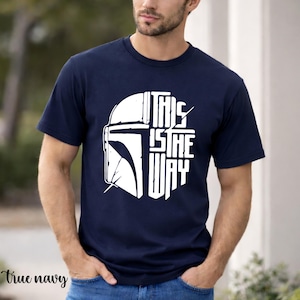 May include: Navy blue t-shirt with a white graphic of a helmet and the text "THIS IS THE WAY". The shirt is short-sleeved and made of a soft material.