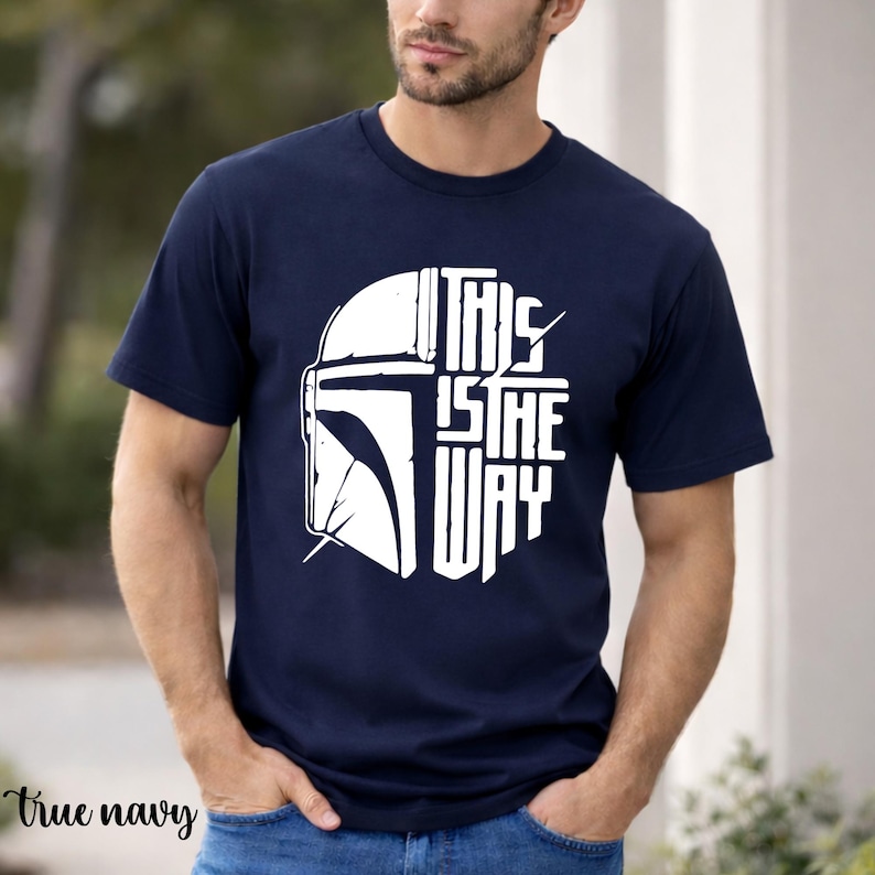 May include: Navy blue t-shirt with a white graphic of a helmet and the text "THIS IS THE WAY". The shirt is short-sleeved and made of a soft material.