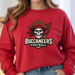 May include: A red sweatshirt featuring a pirate skull wearing a red hat with gold trim and a black eye patch. The words "BUCCANEERS FOOTBALL" are printed below the skull, with a football graphic. The sweatshirt is a solid red color.