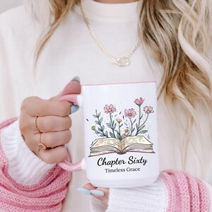 May include: White ceramic mug with a pink handle and rim, featuring a floral design with pink flowers and green leaves. The mug has the text "Chapter Sixty" and "Timeless Grace".