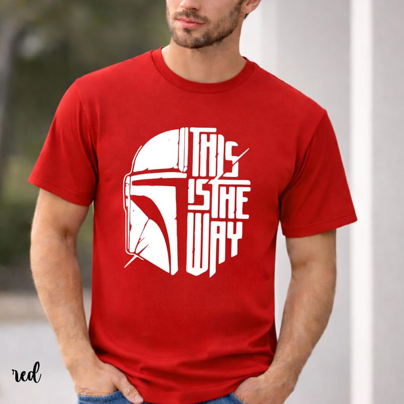May include: Red t-shirt featuring a white graphic of a helmet and the text "THIS IS THE WAY". The design is centered on the front of the shirt. The shirt has short sleeves and a crew neck.