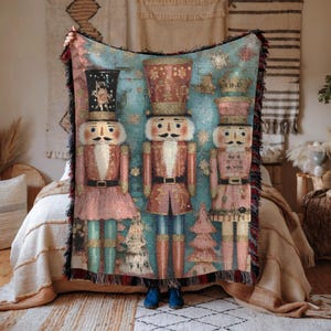 May include: A woven throw blanket featuring three nutcracker figures in red, pink, and blue. The blanket has a fringed edge and a light blue background with snowflakes. The nutcrackers wear tall hats and have mustaches.