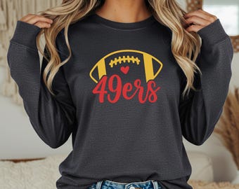 Puede incluir: Una camiseta de manga larga gris oscuro con un gráfico de fútbol americano amarillo y rojo y el texto "49ers". La camiseta tiene cuello redondo y está hecha de un material suave. La palabra "pepper" está impresa en la esquina inferior izquierda.