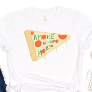May include: White t-shirt with a graphic of a pizza slice with the text "Amore al Primo Morso" printed on it. The shirt is paired with blue jeans and white striped slip-on shoes.